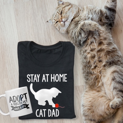 Very cute cat laying next to a black t-shirt that reads: Stay at home Cat Dad and a mug with an Adoption design.