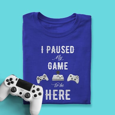 Game controller beside a t-shirt with custom design showing an illustration of a game box and controllers with stylized text that reads: I Paused My Game to Be Here.