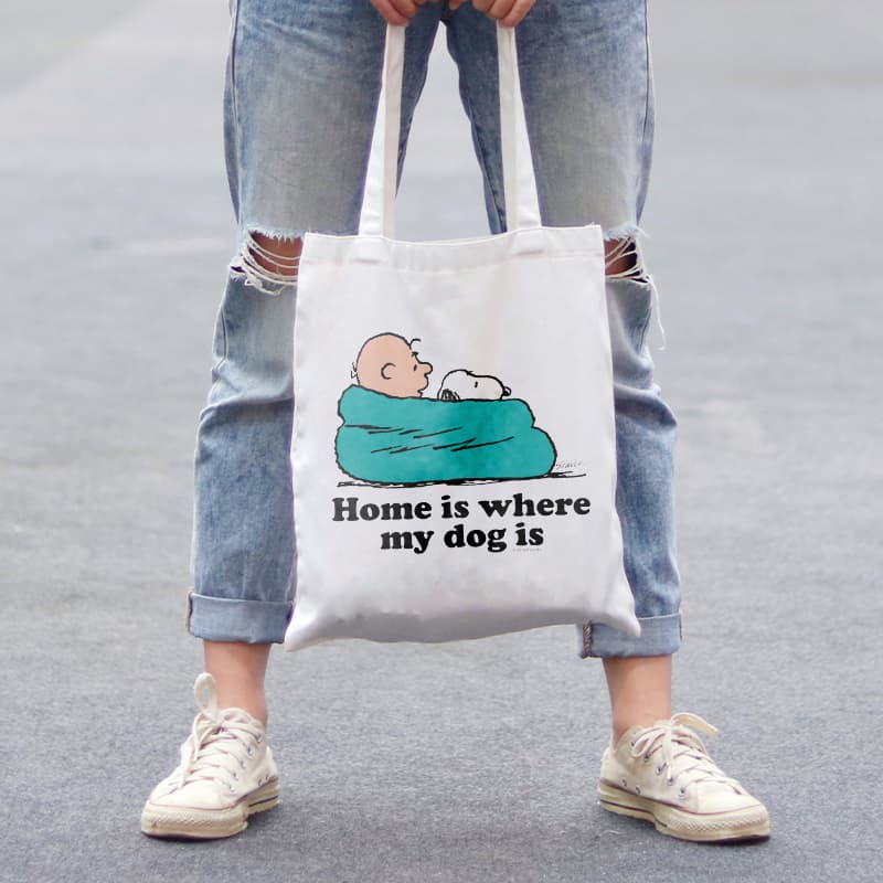 Woman holding a canvas tote bag with an official licensed PEANUTS® design showing Snoopy and Charlie Brown snuggling in a blanket and stylized text that reads: Home is Where My Dog Is.