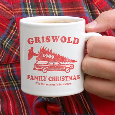 Man in a plaid shirt holding a custom printed mug with an official licensed National Lampoon's Christmas Vacation design showing an illustration of a station wagon with a giant tree on top and stylized text that reads: Griswold Family Christmas 1989.
