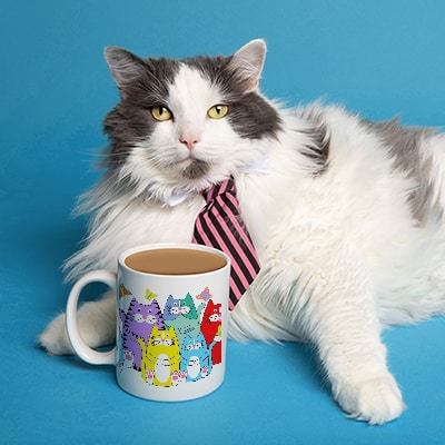 Cute cat wearing a tie sitting beside a coffee mug. The mug has a custom printed design with a funny illustration of a colorful family of cats.