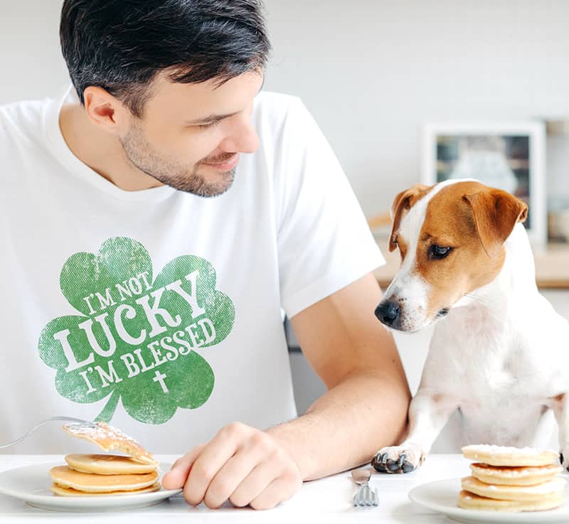 Man sitting at the table eating pancakes with a dog looking hungrily at his plate. Man is wearing a custom printed t-shirt that reads - I'm not lucky, I'm blessed. Contained within a clover.