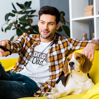 Man sitting on couch with his dog. Man is wearing a custom design t-shirt with stylized text design that reads: Dog Dad.