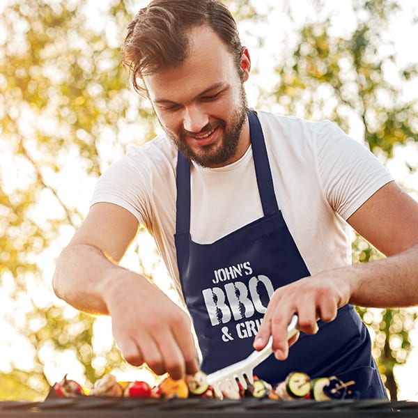 A man grilling wearing a navy apron with a design that reads John's BBQ and Grill