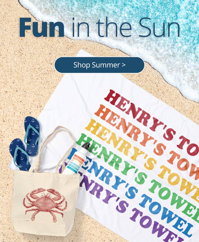 A beach scene with a custom printed towel, water bottle, flip flops, and tote bag all laying on the sand near the water as it washes up on shore.