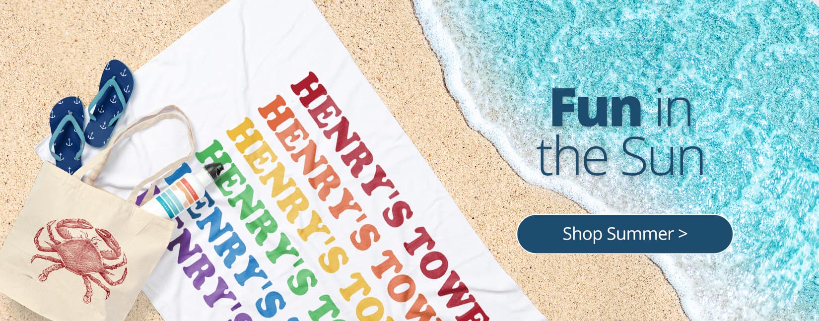 A beach scene with a custom printed towel, water bottle, flip flops, and tote bag all laying on the sand near the water as it washes up on shore.