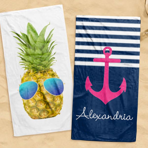 Two beach towels laying on the sand. One has a design of a pineapple with sunglasses and the other is a pink anchor of a blue and white color block with a personalized name on the bottom reading Alexandria.