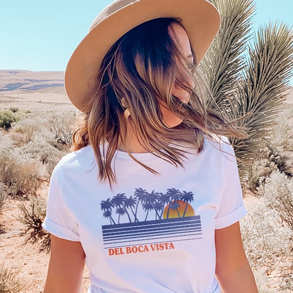 A woman with her hair blowing in the wind wearing a custom printed tee with a Seinfeld design of a sunset behind illustrated palm trees with the words Del Boca Vista underneath it.