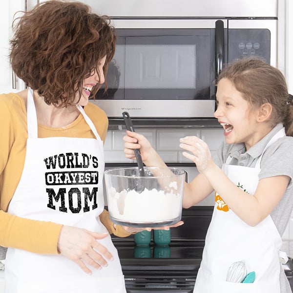 A mother and daughter baking in the kitchen and the mom's apron has a design that reads - World's Okayest Mom.