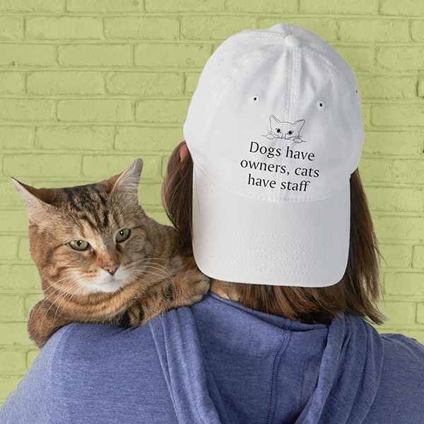 A female wearing a backwards hat with a design that reads - Dogs have owners, cats have staff - while holding an adorable cat on her shoulder.