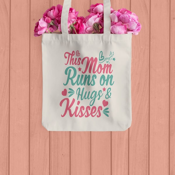 A canvas tote bag with flowers coming out of the top laying on a wooden table with a stylized text design that reads - This Mom Runs on Hugs and Kisses.