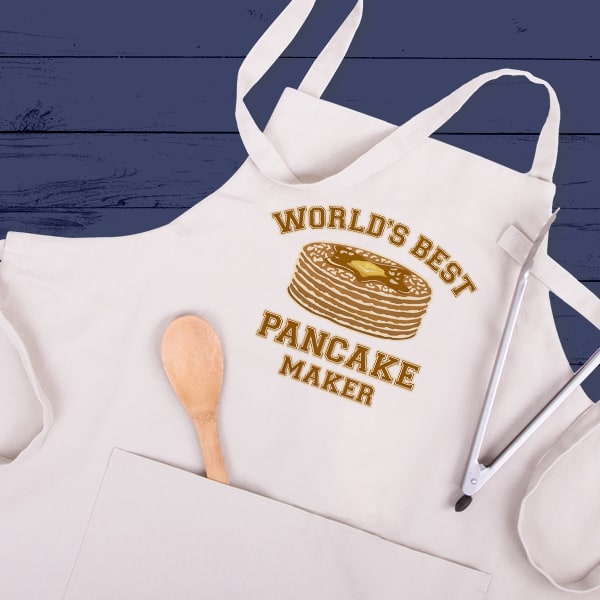 An apron laying on a blue wooden table with a design that reads World's Best Pancake Maker with an illustration of a stack of pancakes.