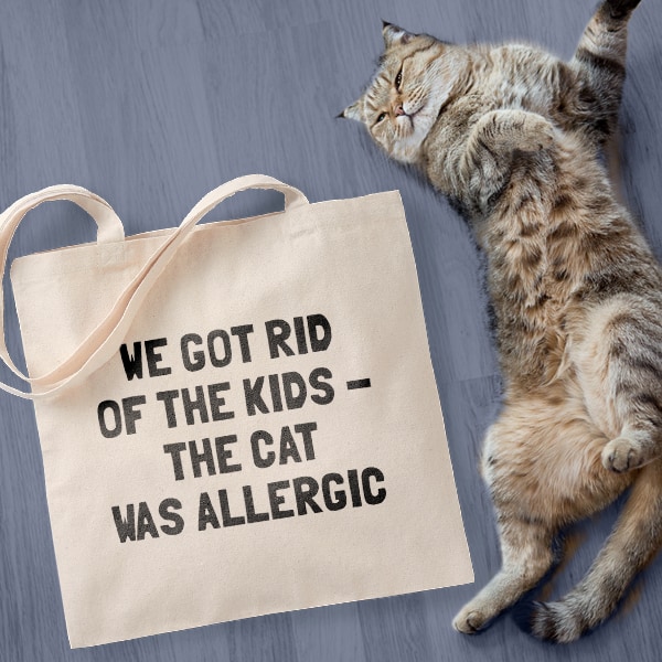 World's cutest cat laying next to a canvas tote bag with a design printed on it that reads We got rid of the kids, the cat was allergic.