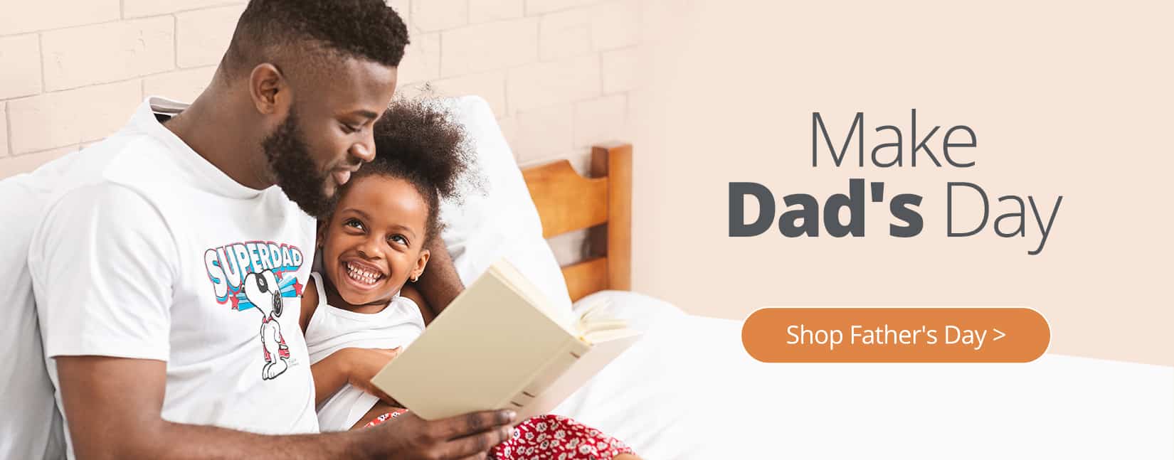 A father in daughter sitting in bed together reading a book. The dad is wearing an officially licensed PEANUTS(R) design on a classic t-shirt. The design shows Snoopy(TM) in a super hero outfit with the word Superdad over him.