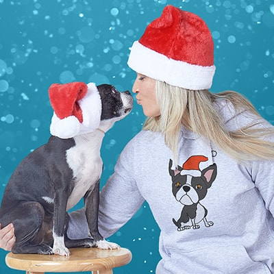 Adorable dog being kissed by its owner while both are wearing a Santa Claus hat. Female owner wearing a hoodie with an illustration of her dog.