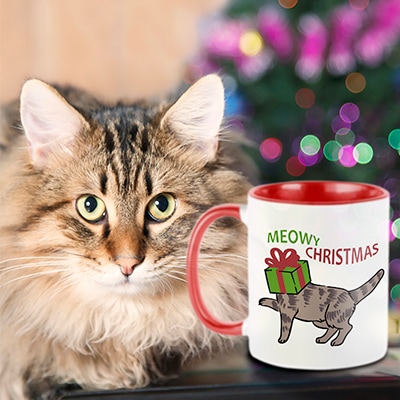 Adorbale fuzzy cat sitting next to a CafePress red handle mug with the design Meowy Christmas on it and an illustration of a silly cat with their head stuck in a gift box.