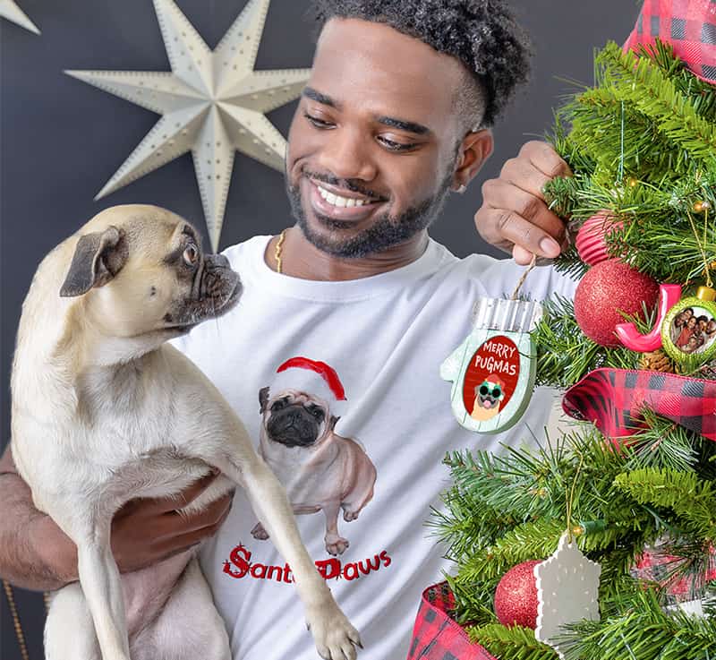 An owner and his pug hanging a Merry Pugmas customized Christmas ornament on his tree, while wearing a custom printed CafePress shirt with an image of his dog in a Santa hat on it reading Santa Paws.
