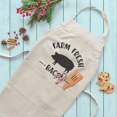 Men's tan apron with custom design with pig silhouette and stylized text reading: Farm Fresh Bacon.