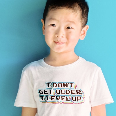 Cute Asian American boy wearing white t-shirt with custom design with gamer stylized text reading: I Don't Get Older, I Level Up.